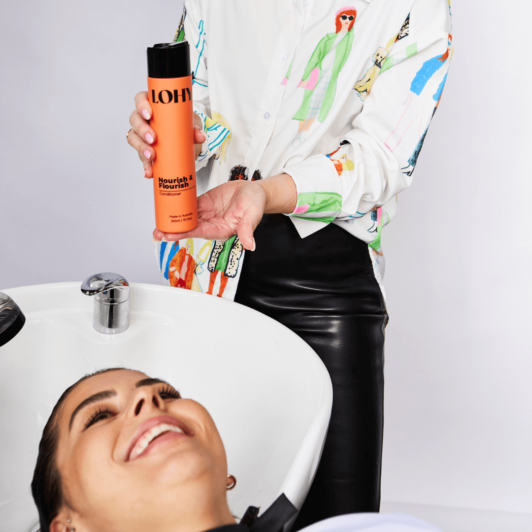 woman holding hair conditioner bottle with woman having hair washed at sink